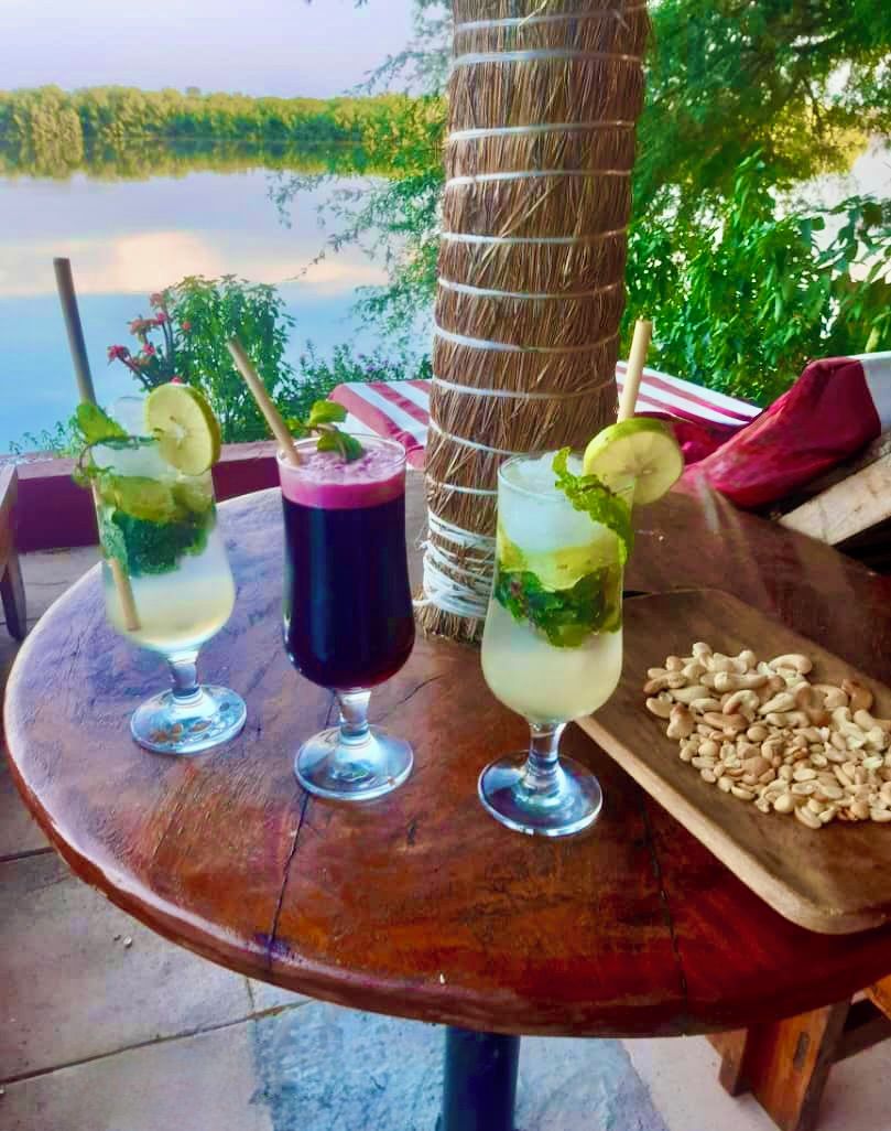 A table with two drinks, a wooden board with nuts, and a view of a lake.