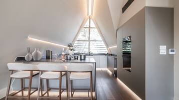 A modern kitchen with a dining area, featuring a long countertop, wooden chairs, and a large window providing natural light.