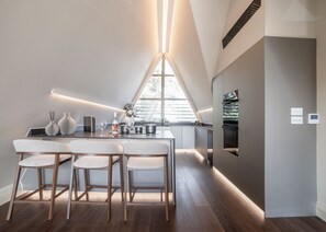 A modern kitchen with a dining area, featuring a long countertop, wooden chairs, and a large window providing natural light.
