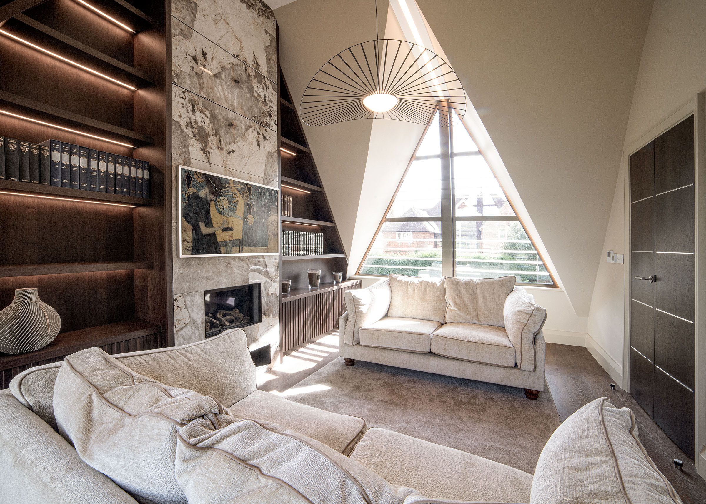 A modern loft bedroom with a sloped ceiling, a built-in bookshelf, a fireplace, a large window, and a cozy seating area.