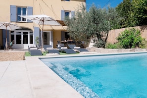 A pool area with lounge chairs and umbrellas, a dining table with chairs, and a view of a building with windows and shutters. - Le Mas de Menard (Barbentane)