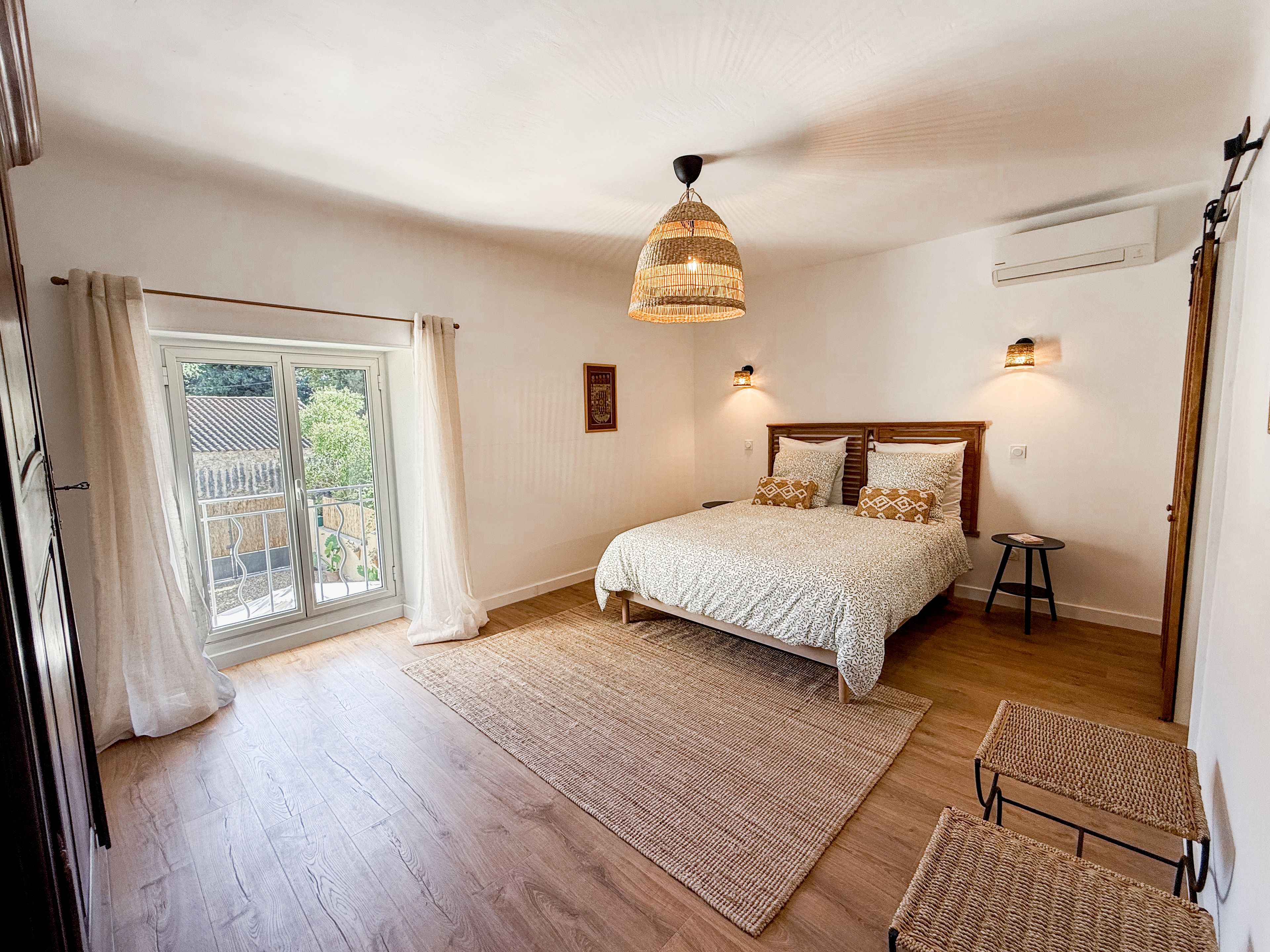 Une chambre à coucher avec un lit, une fenêtre avec des rideaux, un luminaire suspendu et un petit tabouret.