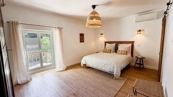 Une chambre à coucher avec un lit, une fenêtre avec des rideaux, un luminaire suspendu et un petit tabouret.