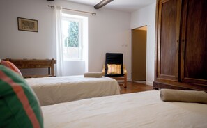 A bedroom with two beds, a wooden wardrobe, a chair with a cushion, and a window with curtains. - Vrbo Property (Barbentane)
