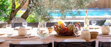 Un'area pranzo all'aperto con un tavolo apparecchiato per due, circondata da ulivi e una piscina sullo sfondo.