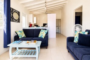 Una sala de estar con un sofá, una mesa de centro y un ventilador. Se observa una cocina al fondo.