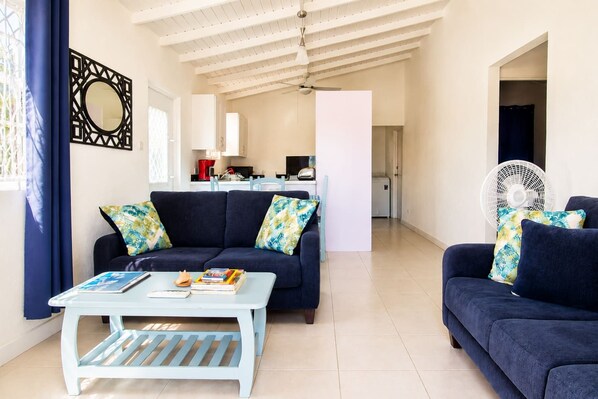 A living room with a sofa, a coffee table, and a fan. There is a kitchen in the background.