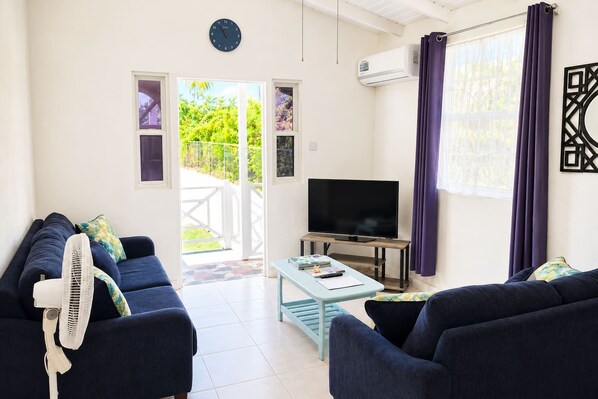 Una sala de estar con televisor, dos sofás, un ventilador y una mesa con libros y controles remotos.