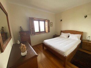 A bedroom with a wooden bed, a dresser, a nightstand, a window, and a mirror. - Casa do Laranjal (Curvos)