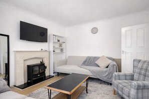 A living room with a fireplace, a sofa, a plaid armchair, a television, and a bookshelf. - 3 Bed House with garden Free Parking near Tottenham Stadium (London)