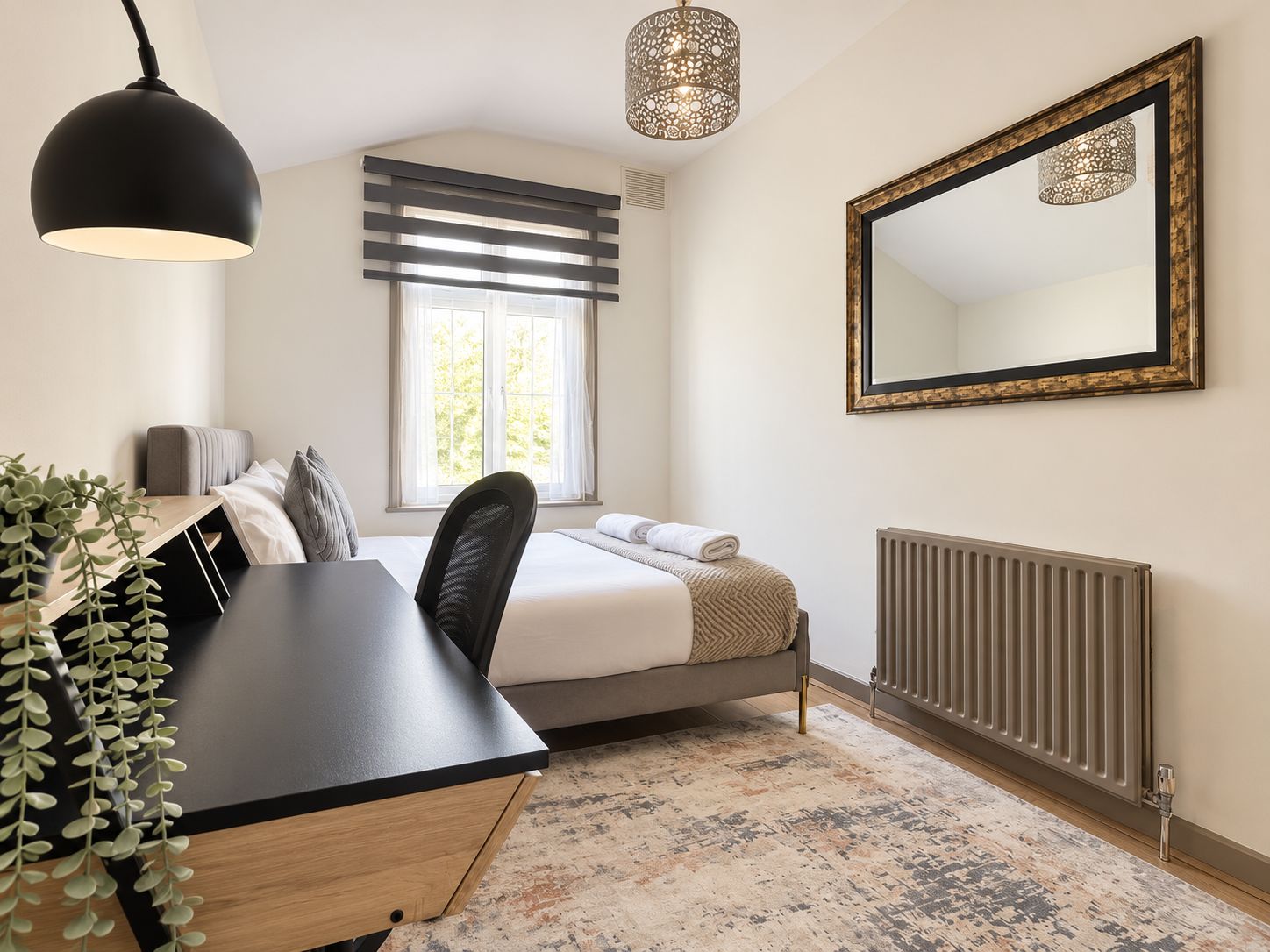 A bedroom with a bed, a desk, a chair, a radiator, a window with blinds, and a hanging light fixture.