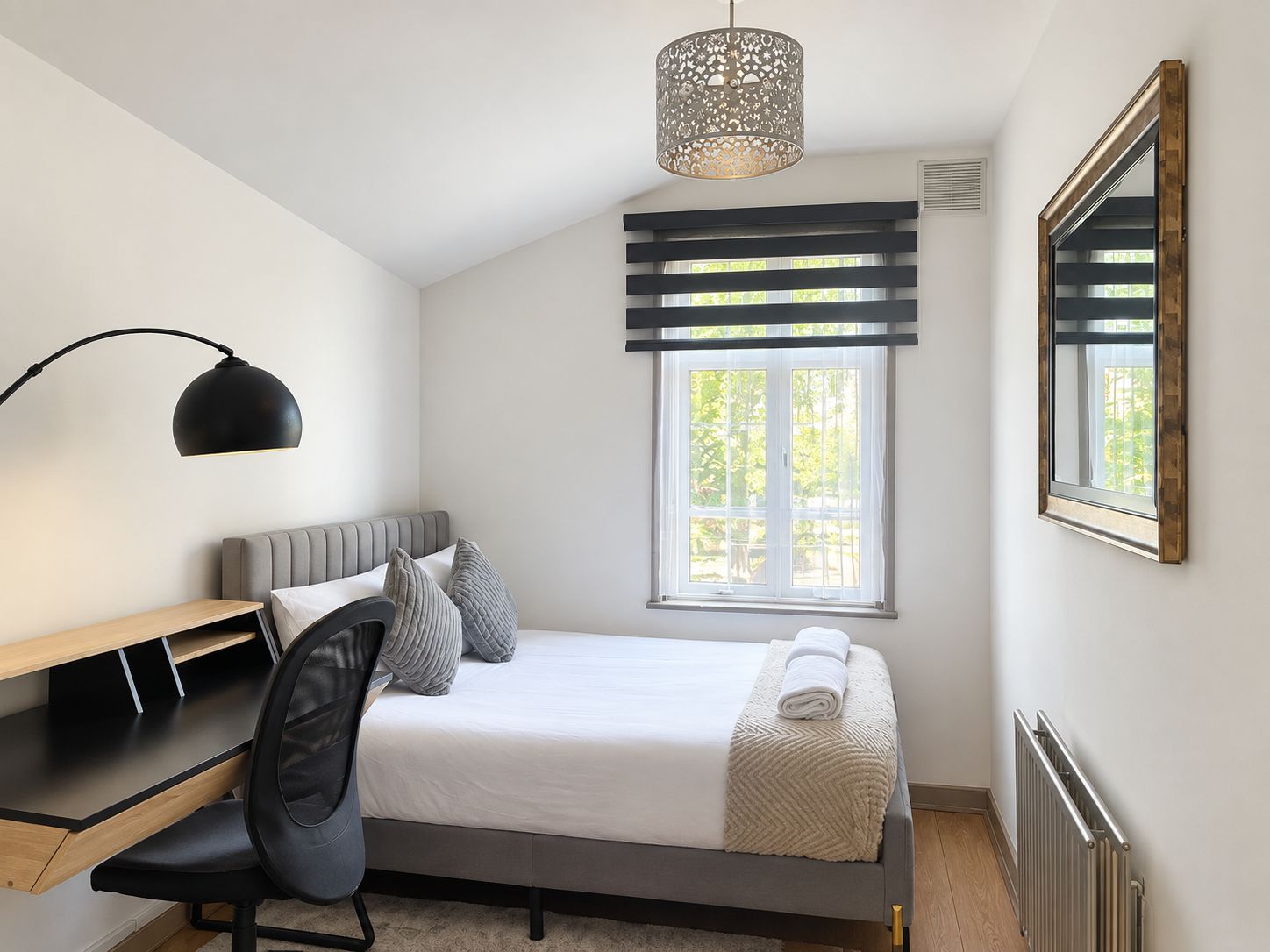Une chambre à coucher comprenant un lit, un bureau, une chaise, une fenêtre avec des stores et un luminaire suspendu.