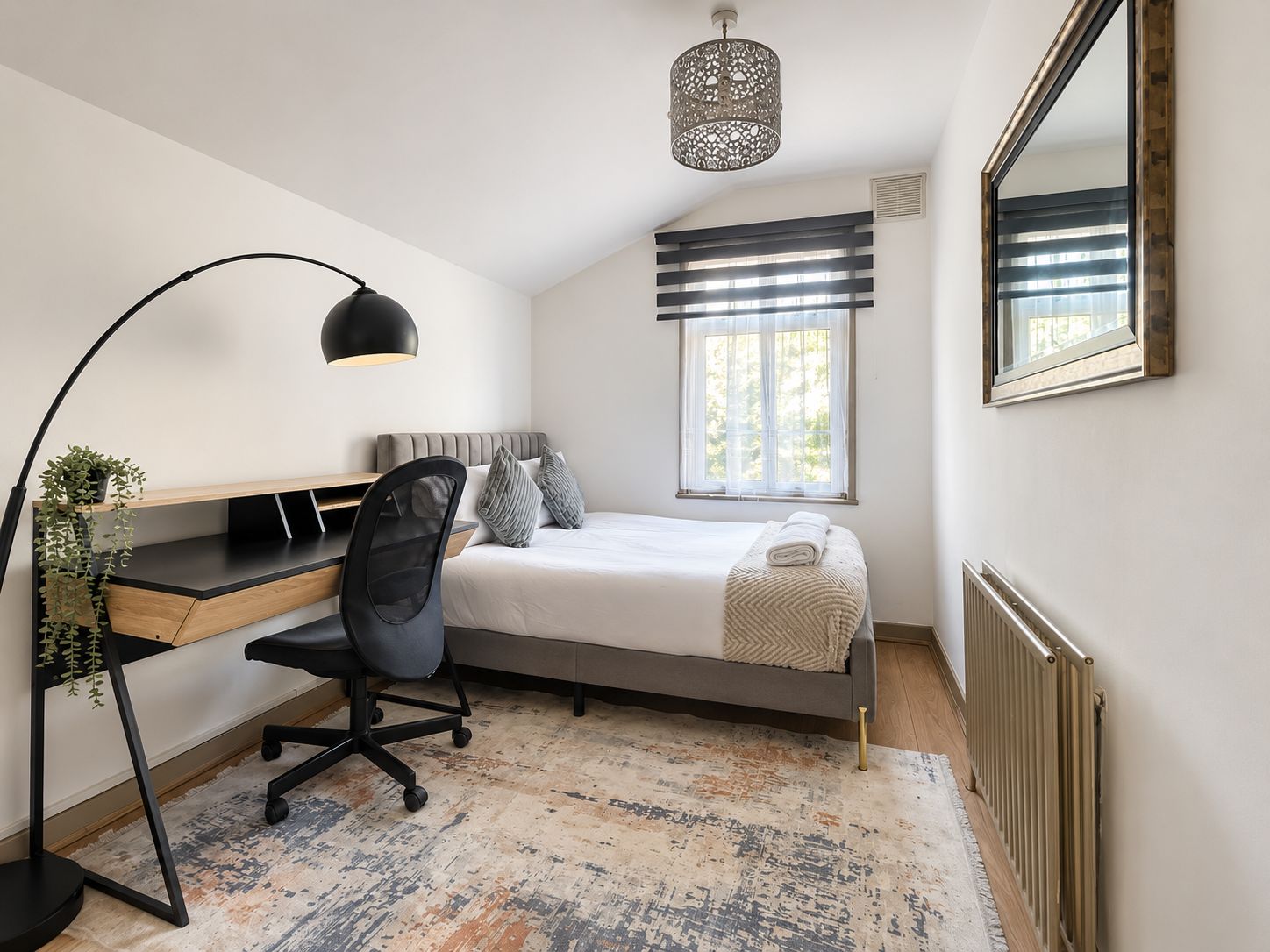 A bedroom with a bed, desk, chair, lamp, window, and radiator.