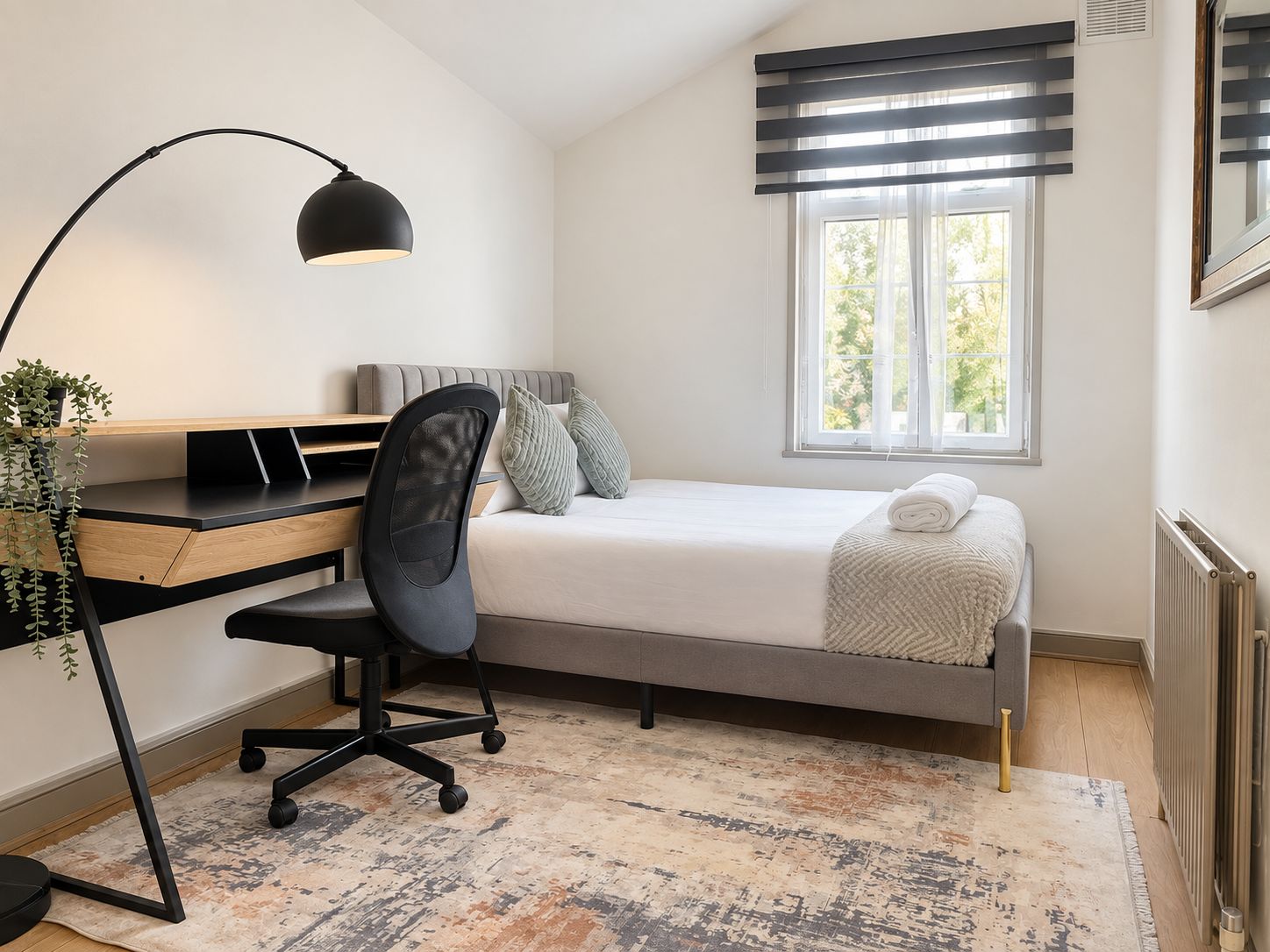 Une chambre à coucher avec un lit, un bureau avec une chaise, une fenêtre avec des stores et un radiateur.