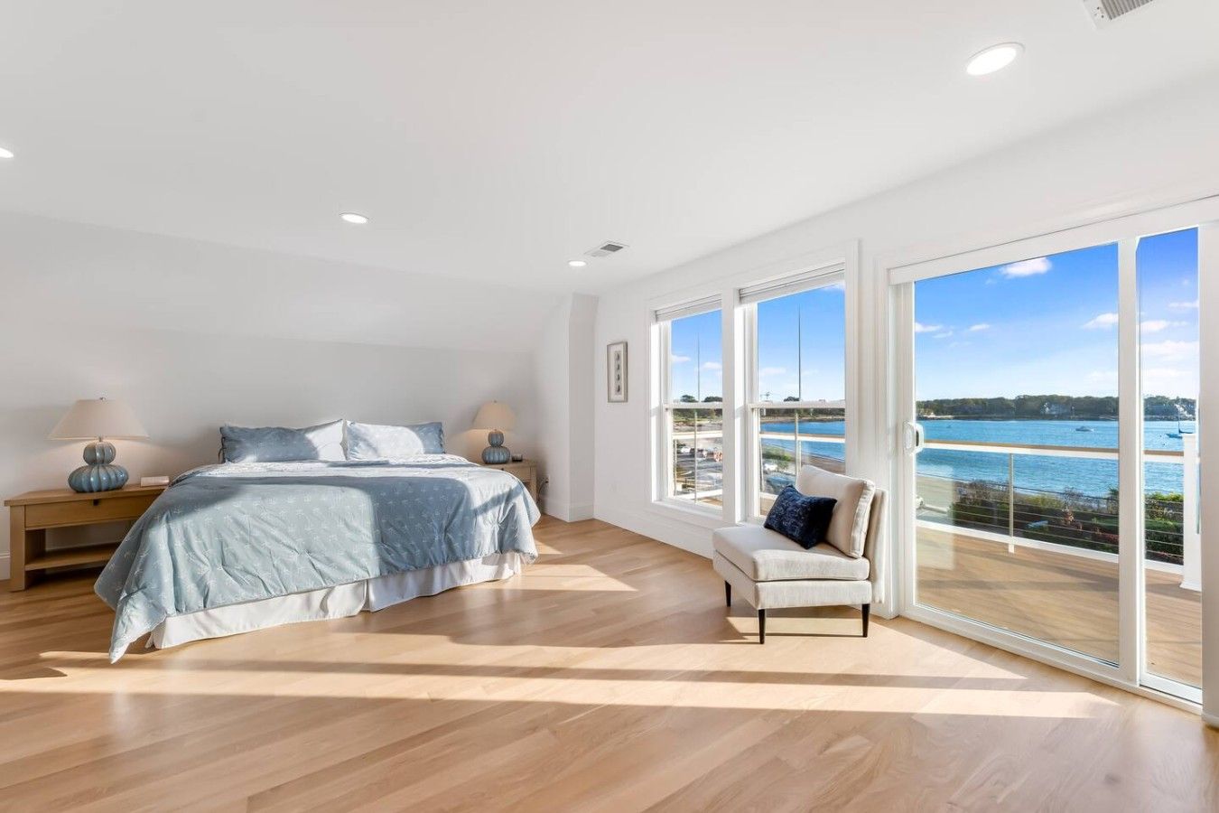 Un dormitorio con una cama grande, una mesita de noche, una silla y vistas al mar a través de amplios ventanales.