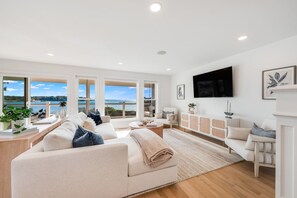 A spacious living room with a large window overlooking a body of water, a flat-screen TV mounted on the wall, and a white fireplace. - Luxury Portside Townhome Steps to Niles Beach (Gloucester)