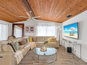 Un salon avec un plafond en bois, une table basse en verre, un canapé beige et une décoration murale sur le thème de la plage.