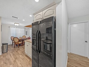 A compact kitchen with a black refrigerator, wooden cabinets, and a dining area with chairs and a table. - Deerfield Coastal House - Cozy Stay (Deerfield Beach)