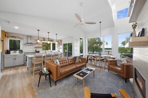 A spacious living area with a large brown leather sofa, a dining table, and chairs. There is a kitchen area with modern appliances and a large window. - Chapel Cove Waterfront Home near Whitewater Lake (Whitewater)