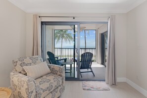 A room with a sofa, a chair, a small table, and a view of the beach through a sliding glass door. - Seaside Condo 109 🌊 Beachfront Condo with Screened Gulf Balcony (Fort Myers Beach)