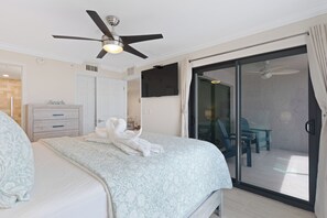 Ein Hotelzimmer mit einem Bett, einem Deckenventilator, einer Glasschiebetür, die auf einen Balkon mit Stühlen führt, und einem an der Wand montierten Fernseher.