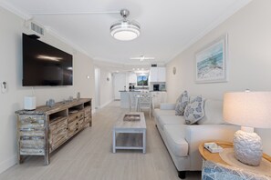 A modern living room with a flat-screen TV, a wooden entertainment unit, a light-colored sofa, and a dining area in the background. - Seaside Condo 109 🌊 Beachfront Condo with Screened Gulf Balcony (Fort Myers Beach)