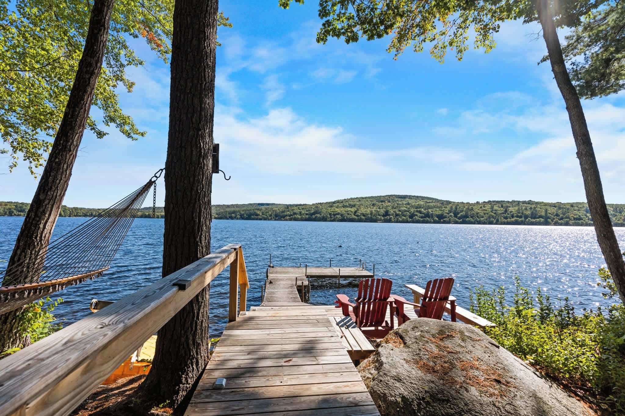 A wooden dock leading to a lake with chairs and a hammock.
