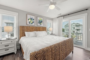 A bedroom with a large bed, two bedside tables with lamps, a ceiling fan, and a view of houses through the window. - Pool, Hot Tub, Rec Room, Fenced Yard, Dogs OK (Corolla)