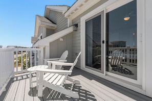 Eine überdachte Terrasse mit weißen Adirondack-Stühlen und einer Glastür, die auf einen Balkon führt.