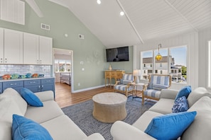 A modern living room with a large sofa, a coffee table, and a television. There is a kitchen area with cabinets and a window. - Private Pool, Hot Tub | Shuffleboard Table | Dogs OK (Corolla)