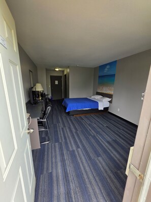 A hotel room with a bed, a desk, a chair, a lamp, and a painting of a beach scene. - Travel Inn resort and Campground (Saskatoon)