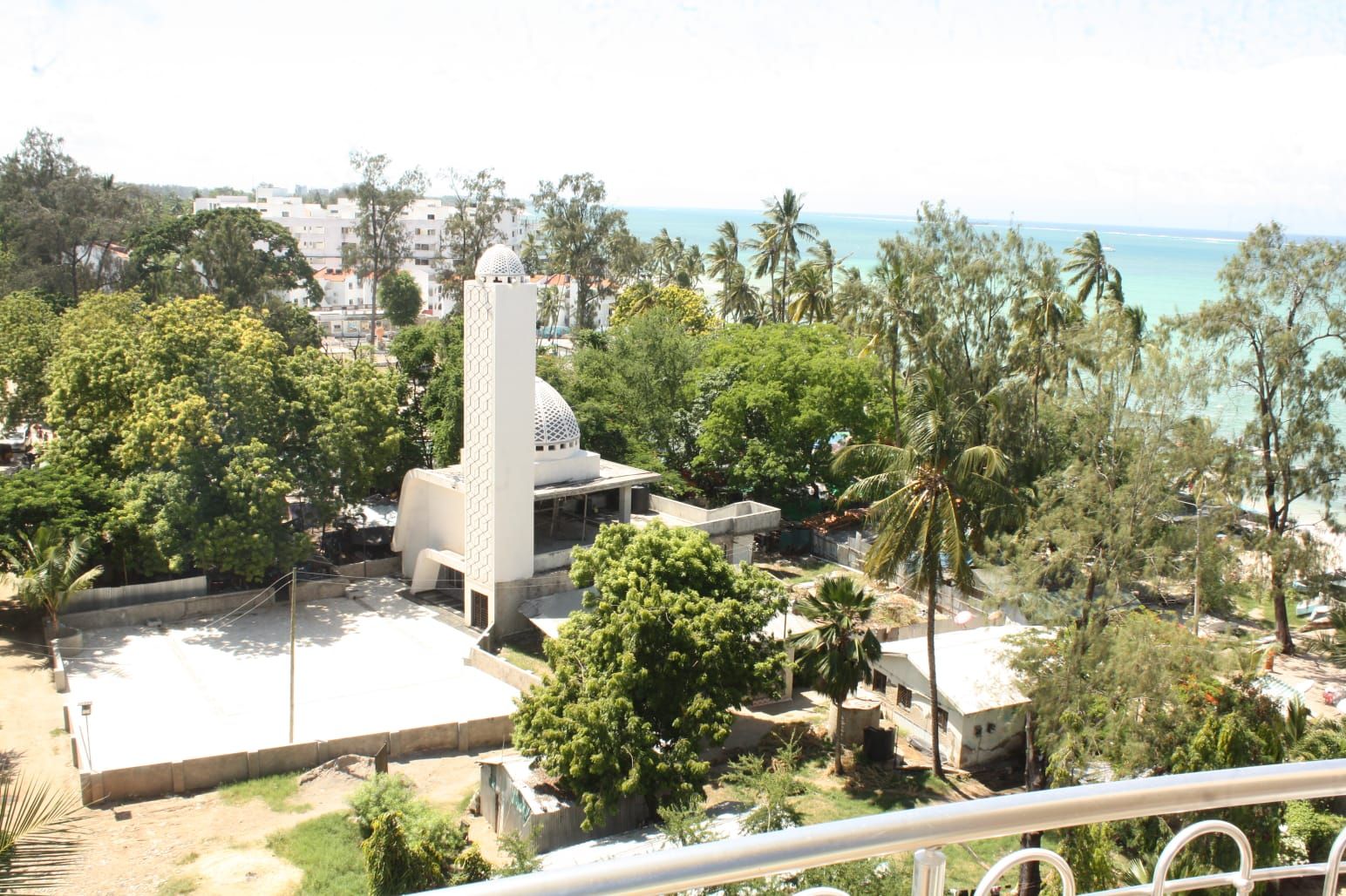 A coastal area with a prominent white building featuring a dome and a tall tower, surrounded by lush greenery and palm trees.