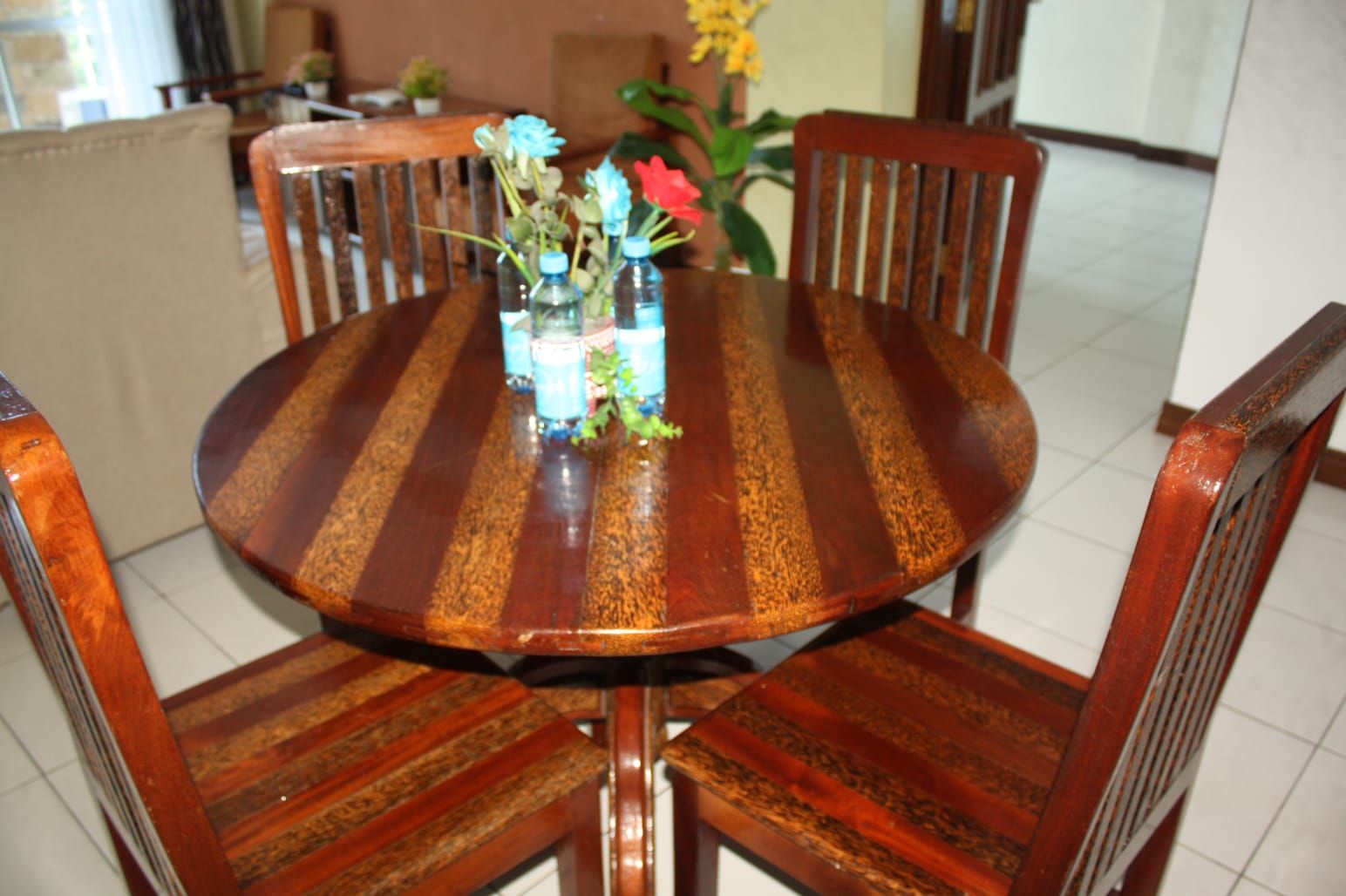 A round wooden dining table with chairs, a vase with flowers, and a bottle of water.
