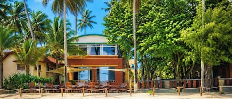 Una propiedad frente a la playa con un edificio de dos pisos, una playa de arena y palmeras.
