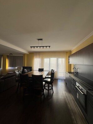 Una cocina moderna con armarios oscuros, un comedor con mesa y sillas de madera, y un ventanal con cortinas translúcidas.