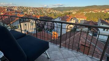Un balcón con una silla lounge negra, barandilla metálica y vista a una zona residencial con casas y colinas al fondo.