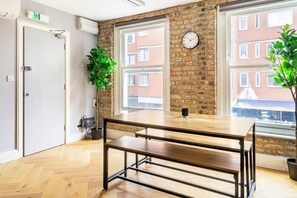 A room with a wooden table, two potted plants, a clock on the wall, and a view of a brick building through a window. - Luxury City Apartment in the Heart of Farringdon (London)