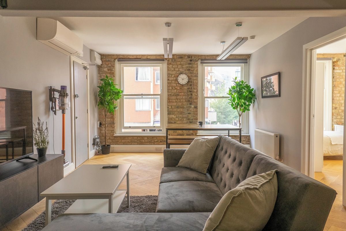 A modern living room with a sofa, coffee table, and a TV on a cabinet. There are two windows with plants, a brick wall, and a clock on the wall.