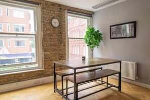 A room with a brick wall, large windows, a wooden table with benches, a clock, and a framed picture on the wall. - Luxury City Apartment in the Heart of Farringdon (London)
