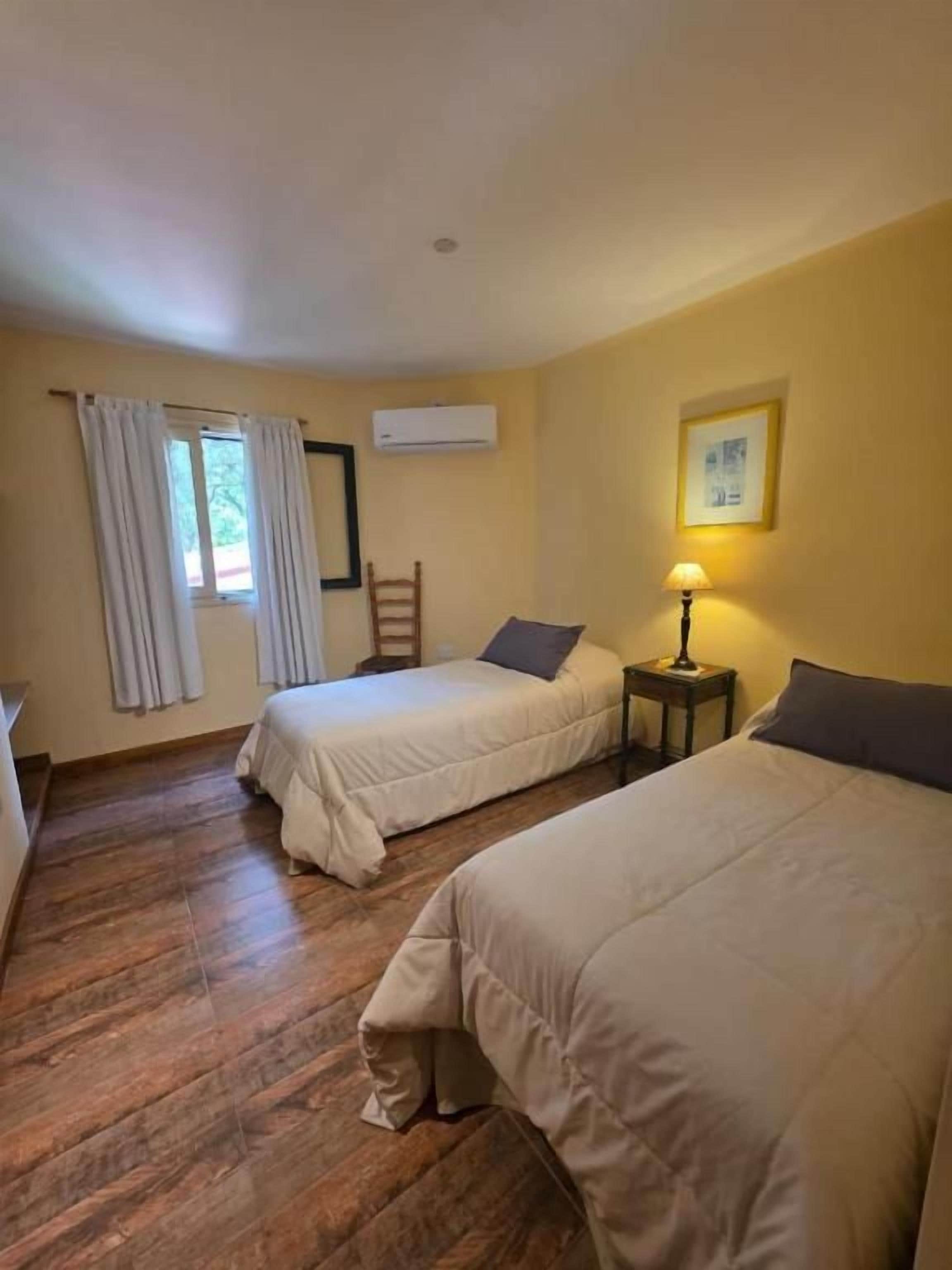 A room with two beds, a window with curtains, a wall-mounted air conditioner, and a small table with a lamp.