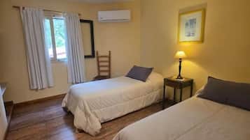 A room with two beds, a window with curtains, a wall-mounted air conditioner, and a small table with a lamp.