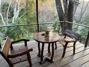 Una terraza de madera con una mesa redonda y dos bancos, con vista a una zona boscosa.