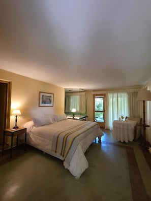 Habitación de hotel con cama, mesitas de noche, ventana con cortinas y una pequeña mesa con lámpara.