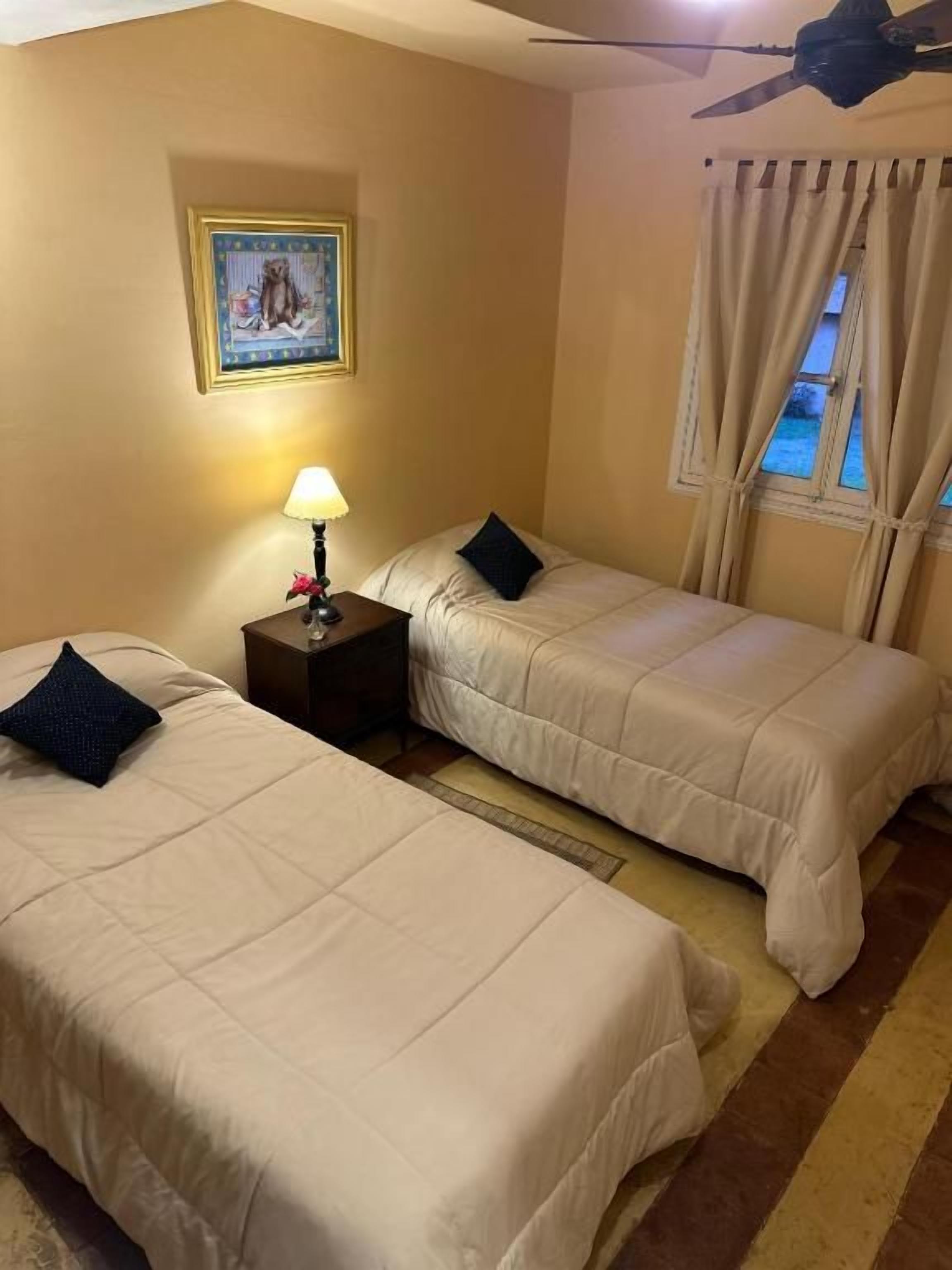 Habitación de hotel con dos camas, una mesita de noche con lámpara, ventilador de techo, ventana con cortinas e imagen colgada en la pared.