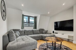 A modern living room with a sectional sofa, a round coffee table, and a flat-screen TV mounted on the wall. - 1BR Flat 1 Min from Finsbury Park Station (London)