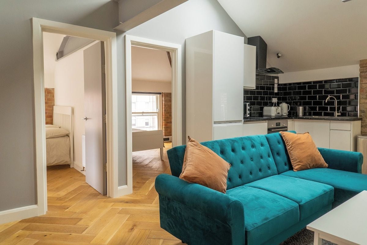 A modern living space with a teal sofa, a white table, and a kitchen area with black and white appliances.