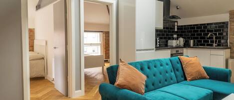 A modern living space with a teal sofa, a white table, and a kitchen area with black and white appliances.