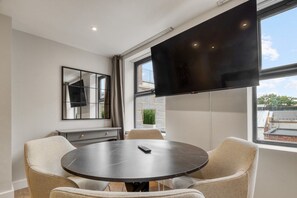 A modern dining area with a round table, chairs, a flat-screen TV mounted on the wall, and a window with a view of buildings. - Deluxe Studio | 1 Minute to Finsbury Park Station (London)