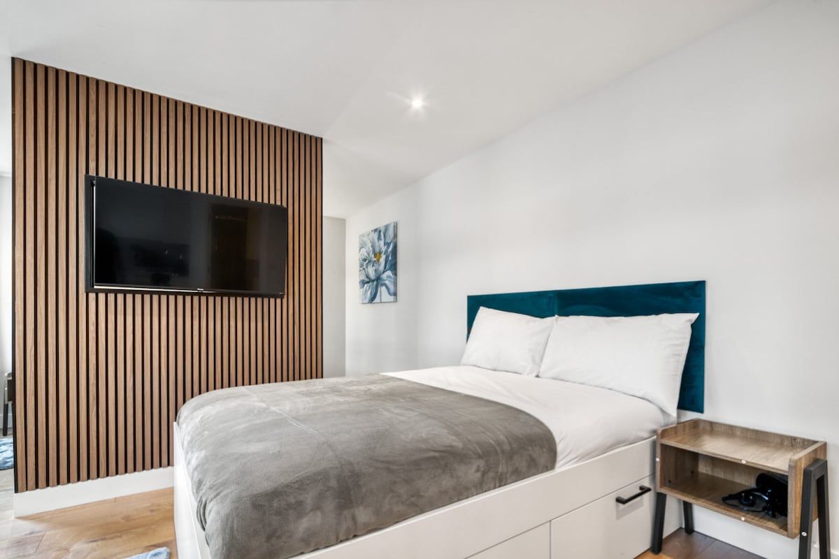 Une chambre moderne avec un lit, un téléviseur fixé sur un mur recouvert de panneaux en bois, une table de chevet et un tableau accroché au mur.