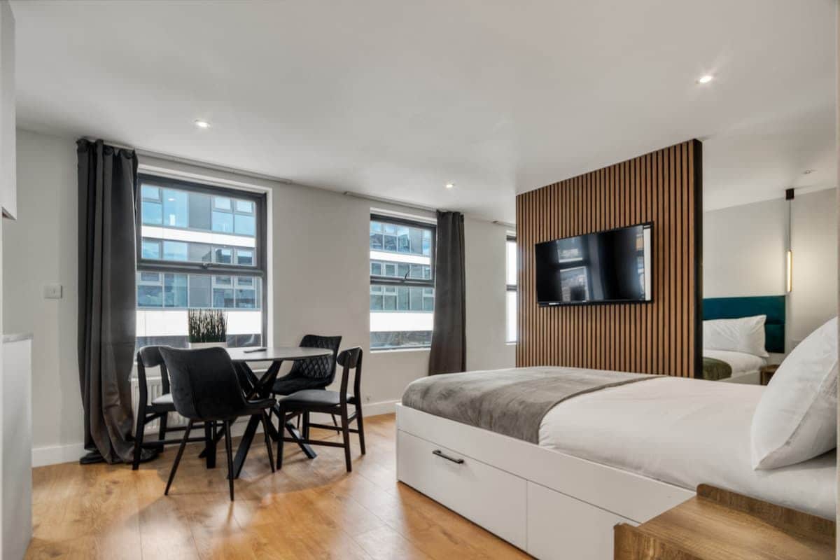Une chambre d’hôtel moderne équipée d’un lit, d’un téléviseur fixé au mur, d’un coin repas avec des chaises et de grandes fenêtres.
